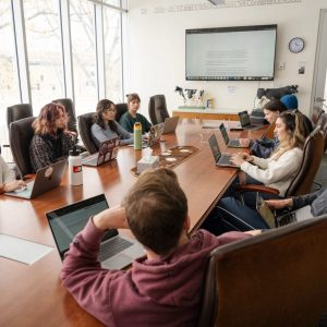 Classroom of students sitting around a conference table, looking at each other and laptops